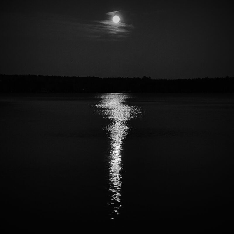 A full moon with a few clouds rises over a calm dark lake, reflecting brightly in the water