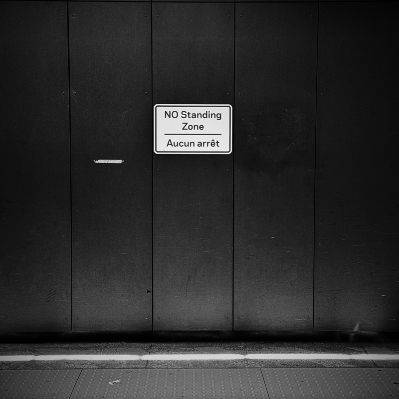 A white sign with red lettering states NO Standing Zone against a plain dark wall