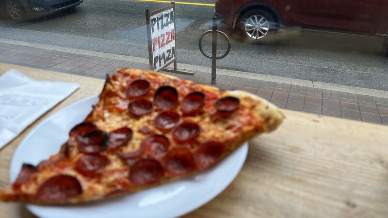 A slice of pizza on a white plate in front of a window overlooking a wet street and a sign saying pizza pizza pizza.