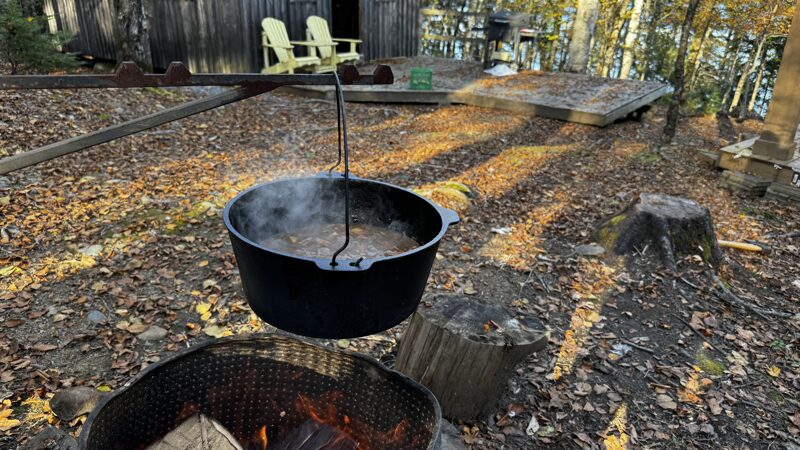 Supper in the woods Beef stew cooks in a dark cast iron pot over a roaring fire outdoors in front of a wood cabin in the woods