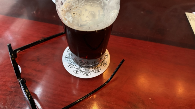 Checked in: Stillwell A pint glass half-filled with dark brown beer and a frothy head sits next to glasses on a dark red bar top
