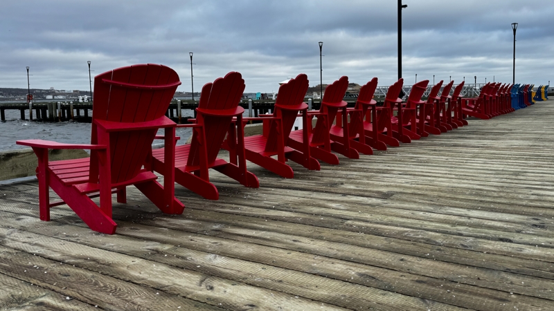A long row of red deck chairs sit perfectly on the harbourfront boardwalk in front of a choppy harbour underneath grey, cloudy skies