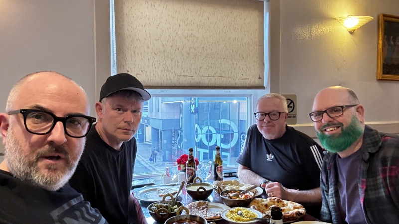Four guys at a table covered in Indian dishes. The background window overlooks a street.