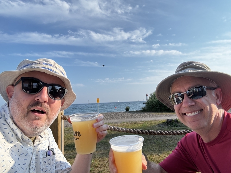 Two middle-aged guys in sun hats with beers sitting at a picnic table on the shores of a huge lake