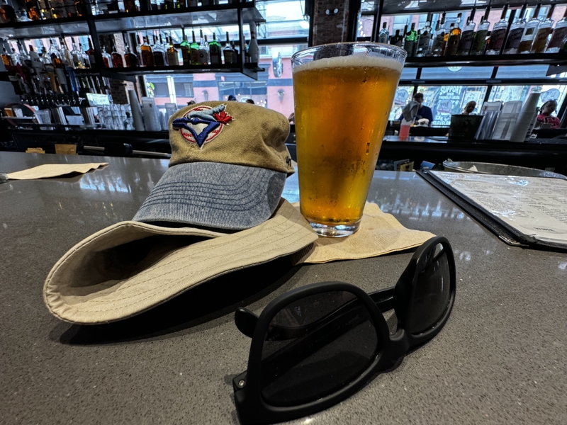 A cold blonde pint of beer sits next to a Toronto Blue Jays hat on a bar top.