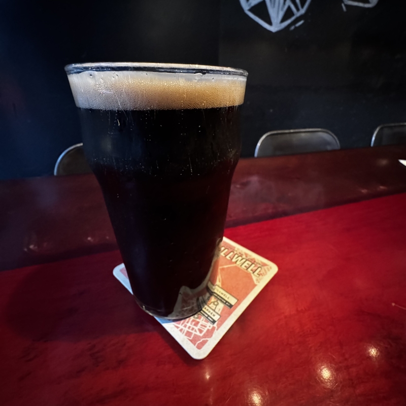 Checked in: Stillwell A pint glass of dark beer sits on a dark red wood table with shiny metal chairs pulled up to it, a blackboard covers the wall behind