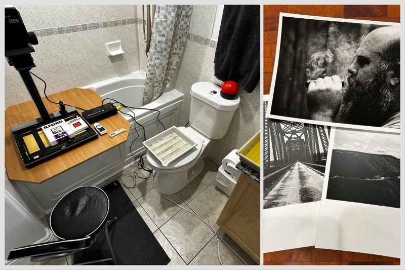 A photo enlarger rests on two boards across a bathtub attached to a digital darkroom timer. A tray of print developer sits on a toilet with a red safelight on the tank.