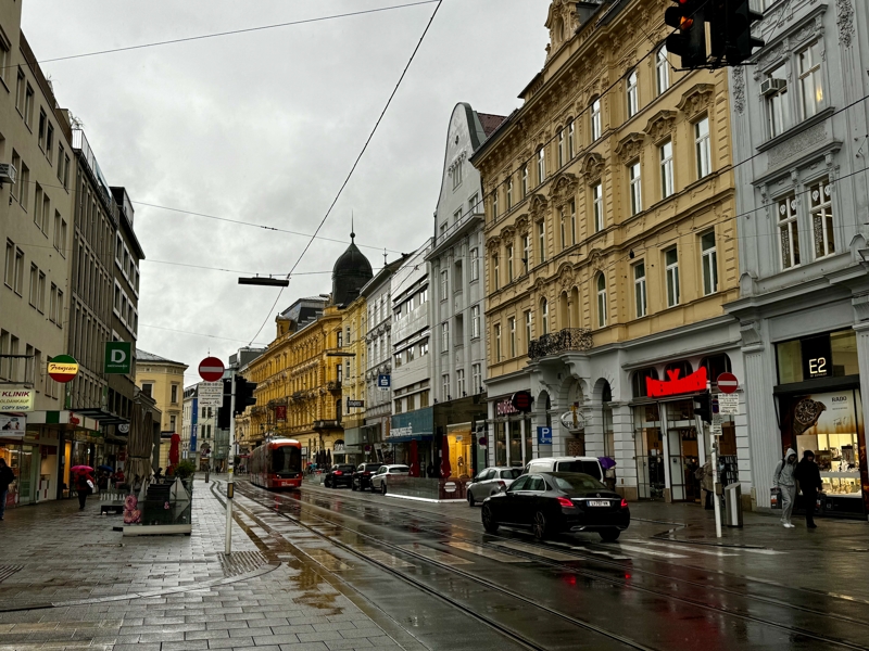 A very rainy and grey Austrian street scene with what would be very colourful buildings and the local tram going by