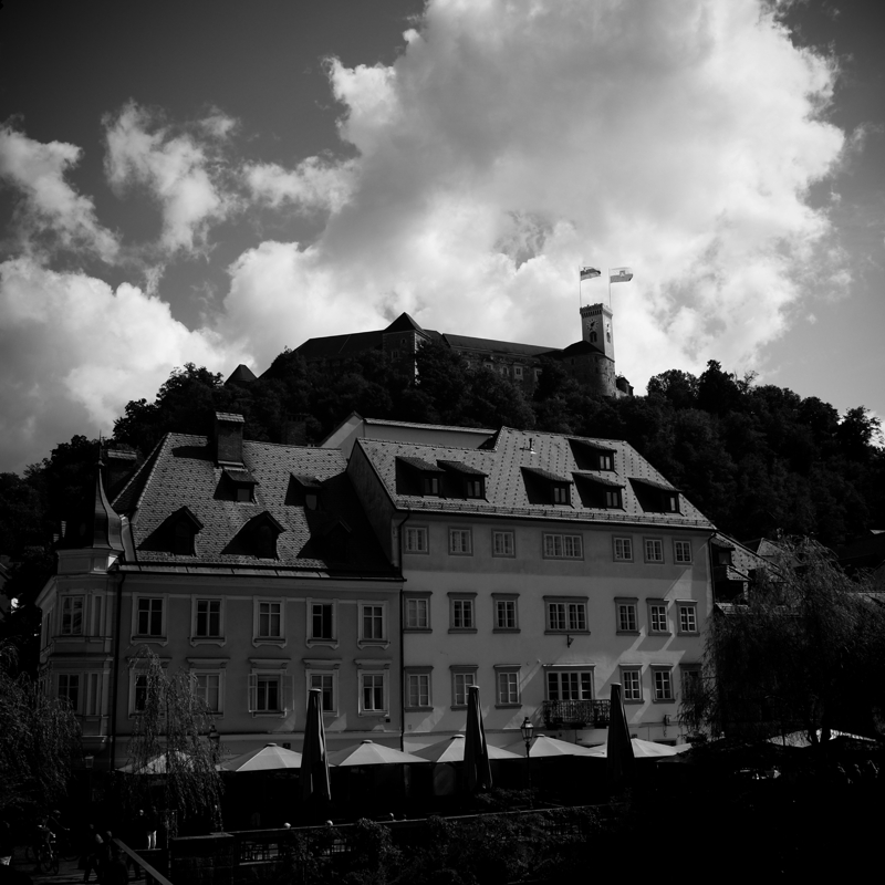 Bright white 19th century buildings are seen against dark trees on the slope of a tall hill with a castle flying the flag of Slovenia
