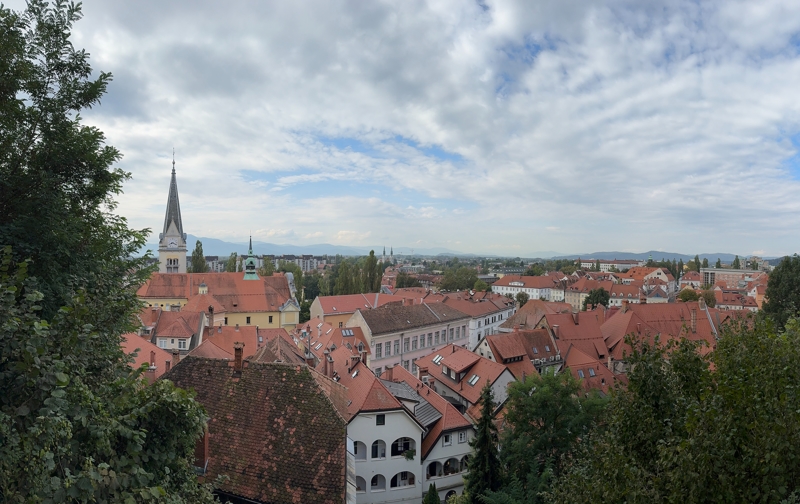 A church spire towers above white buildings with light red roofs with mountains in the distance