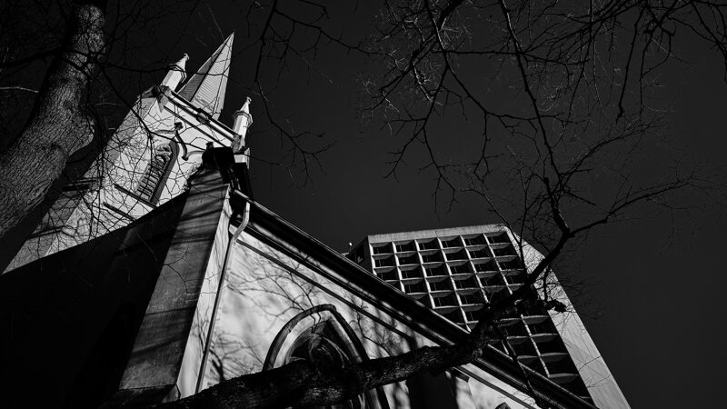 Old and new A brightly lit church is seen set against a dark sky with a modern brightly lit building in the background. Bare trees in the foreground frame the chrurch.