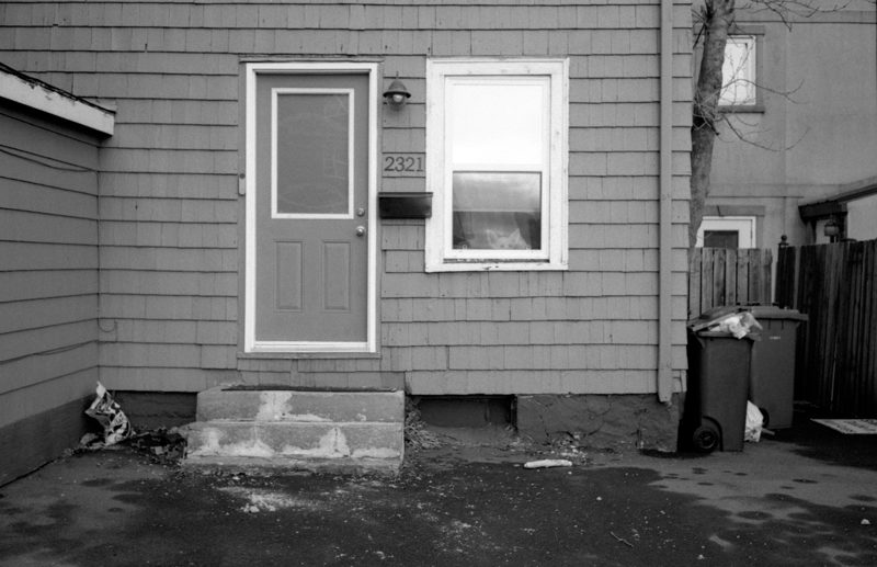 A white dog with ears perked up peers out of a window on the side of an apartment. The building has dark wood siding and a white door with concrete steps leading up to it.