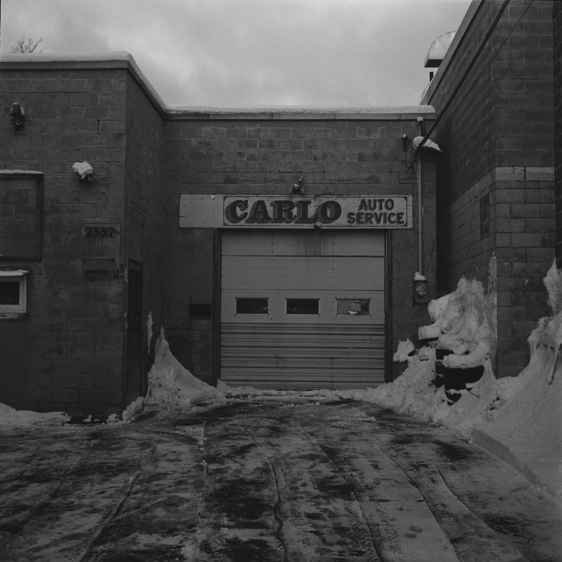 North-end auto service A closed garage door with three small windows in it below a sign reading Carlo Auto Service, there is snow piled up against the grey brick building