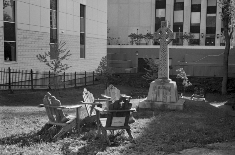 Four wooden chairs with crosses cut out of the back sit in a group on a lawn. There is a large Celtic cross monument next to them while buildings surround the lawn