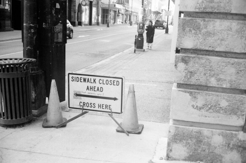 That means you A white sign with the text sidewalk closed ahead cross here and a handwritten this means you is seen blocking a sidewalk in Halifax. A pedestrian can be seen in the background reading posters on a pole.