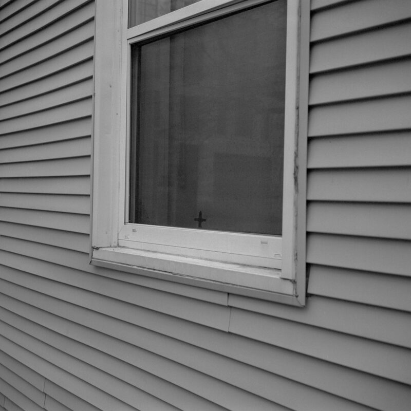 A small, dark cross sits in a closed window, the curtain behind it is light, dark lines of vinyl siding cross horizontally