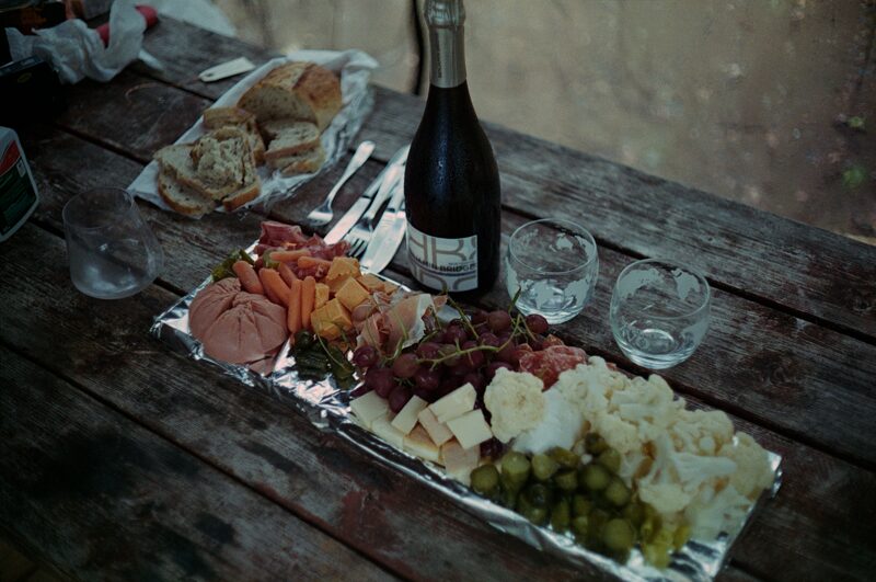 A meal of charcuterie is placed on a dark picnic table. There is a bottle of sparkling wine alongside the food.