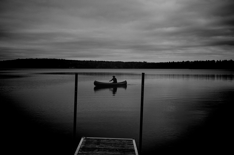 A person sits in a canoe slightly away from shore on a calm lake under cloudy skies, there are two posts at the end of a small wharf at the bottom of the scene