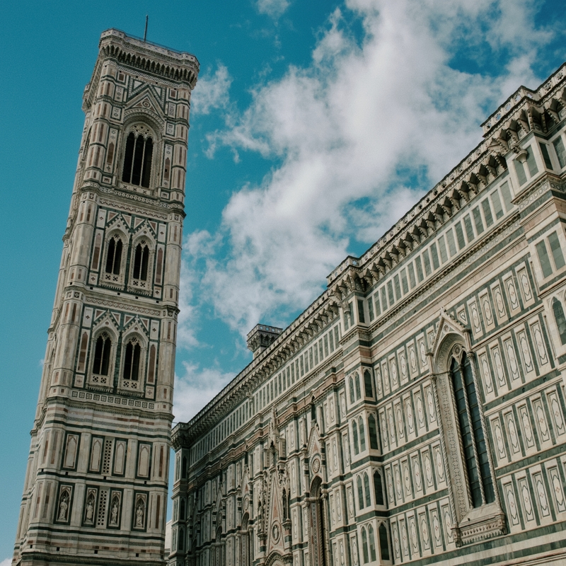 Cathedral of Santa Maria del Fiore, September, 2024 A marble-covered church and separate bell tower curve up toward each other against a bright blue sky and fluffy white clouds