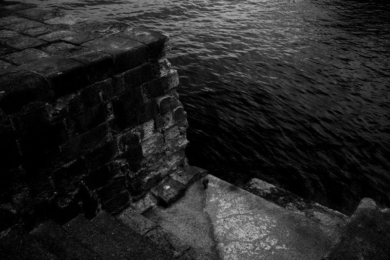 Wharf meets water, Flores Island, May 2025 Dark stones made of volcanic rocks are made into a wharf, steps lead down to the dark water, an iron ring rusts on a step