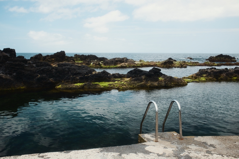 The top of a shiny metal swimming pool ladder is attached to a grey concrete platform overlooking clear cold water with black volcanic rock and the Atlantic Ocean beyond