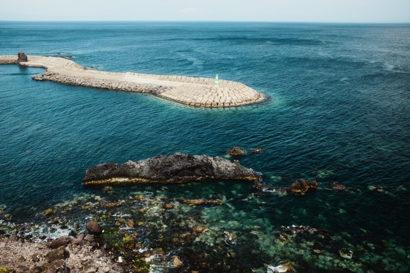 Farolim do Porto das Poças, May 2025 A small green and white striped lighthouse stands alone at the end of a concrete breakwater out in a blue-green ocean with rocks breaking the surface