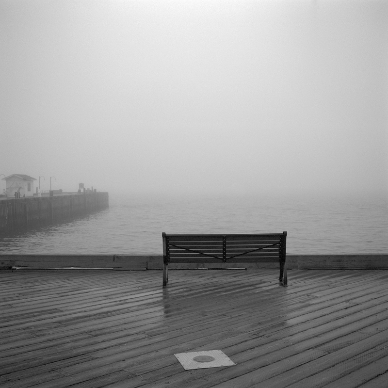 Into the fog An empty wooden bench is seen from behind looking out towards a fog-covered harbour. A wharf with a small white shack and lampposts extends out into the water