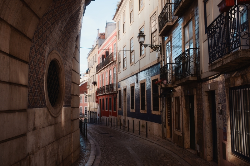 A narrow cobblestone street curves to the left, lined with colourfully painted buildings and a low sun shining on the upper portions