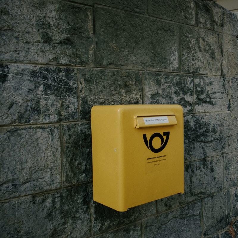 More love letters please! A small bright yellow mailbox is attached to a grey stone wall, there is a small white sticker reading More letters please! stick to the slot