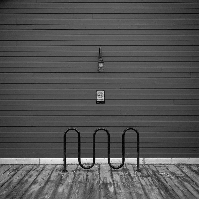 Utilitarian alignment Against the backdrop of a building with horizontal siding a marine light, no smoking sign, and wavy black bike rack line up