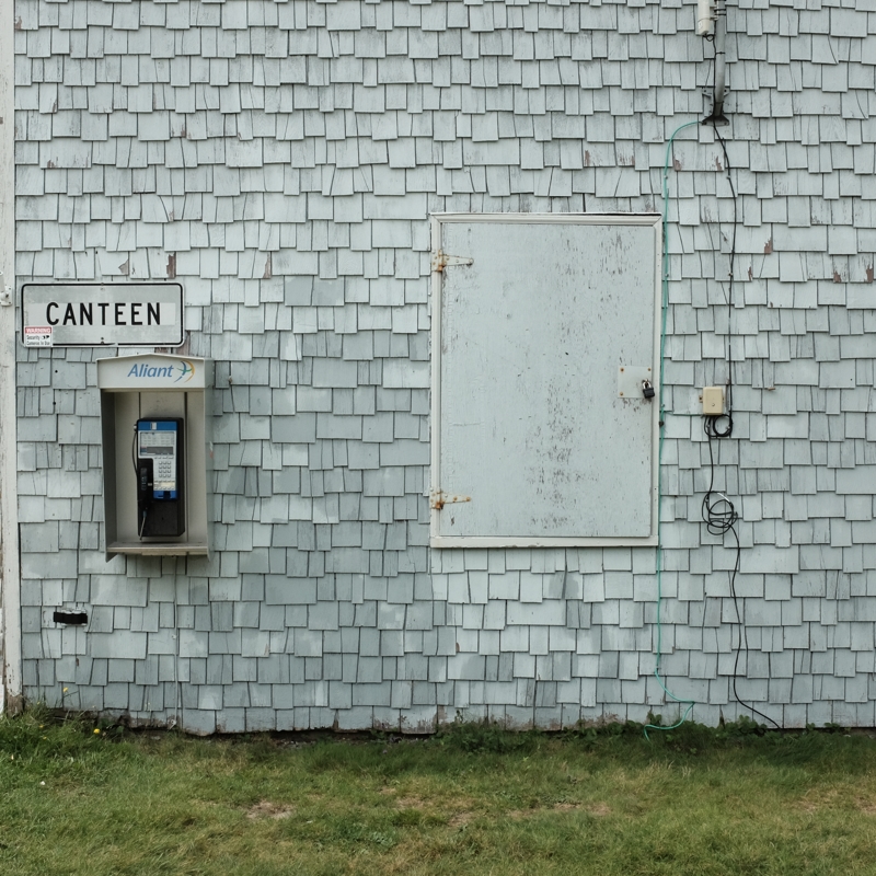 A weathered, very light blue building with an old but working phone attached to the wall underneath a black-and-white sign that reads CANTEEN