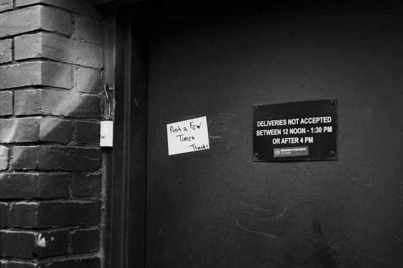 Just a few times A dark metal door has two signs attached, one with delivery hours, the other with instructions to give the for a push a few times