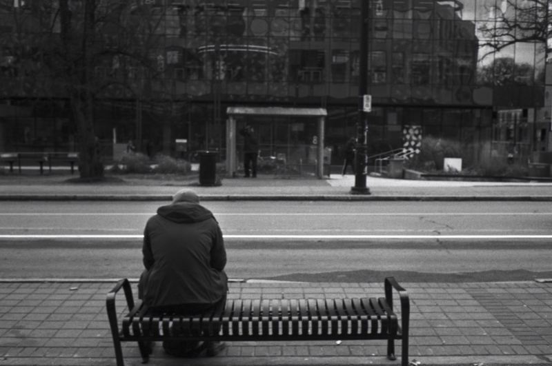 Waiting, November 2024 A person sits alone on a black metal city bench next to a street, a building with reflective windows is in the distance