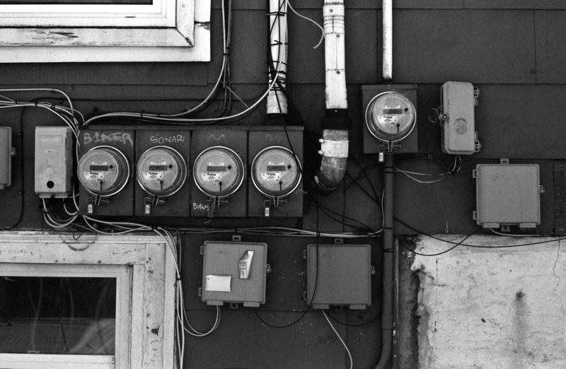 Connections Utility meters and a mess of wires are attached to the side of a house with wood siding. There are wires everywhere, of various colours, some even cut.