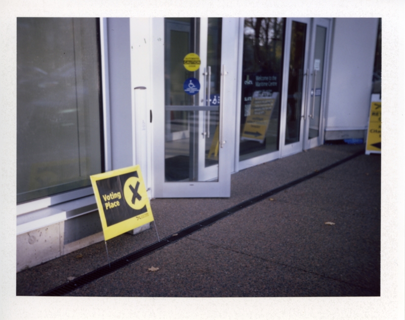 Voting Place, October, 2024 A black and yellow plastic sign rests against a light-coloured wall with a message stating Voting Place